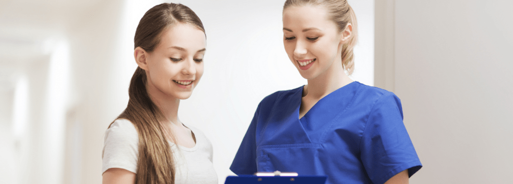 An RMT and their client are standing in a hallway in an indoor setting with white walls and bright lighting. The RMT is holding a clipboard showing the client their health information.