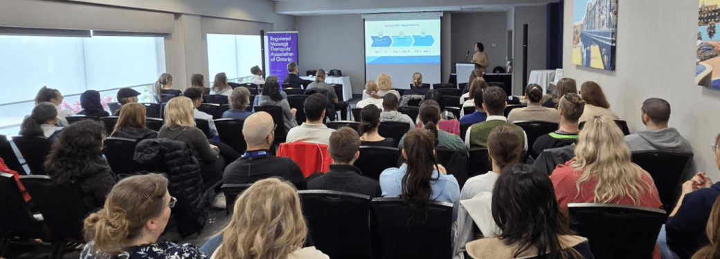 A conference room filled with people seated and facing a presentation screen at the front. The presenter stands near the screen, which displays a slide with diagrams and text. The audience consists of RMTs/MTs attentively watching the presentation. Large windows on one side of the room allow natural light to enter, and a banner near the front reads 'Registered Massage Therapists' Association of Ontario.'