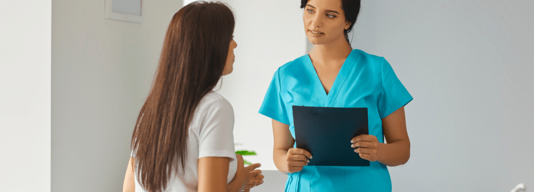 An RMT having a conversation with their client. The client is in a white shirt with long hair and the RMT in blue medical scrubs holding a black clipboard.