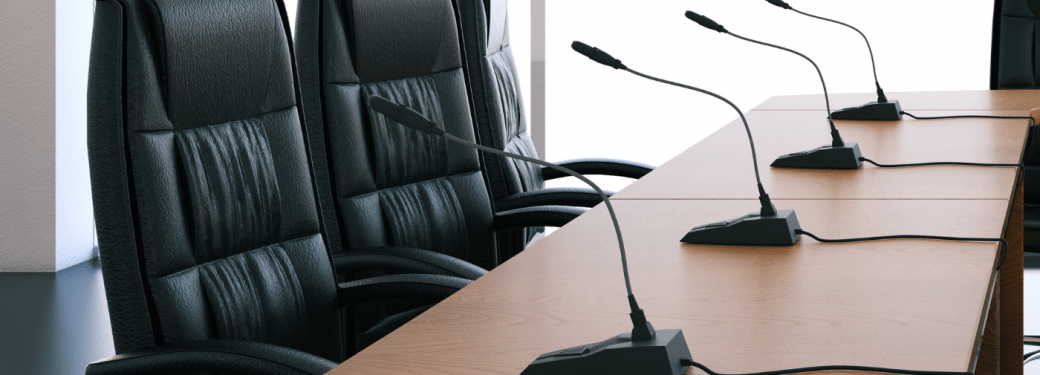 Empty conference room table with microphones and black chairs arranged around.