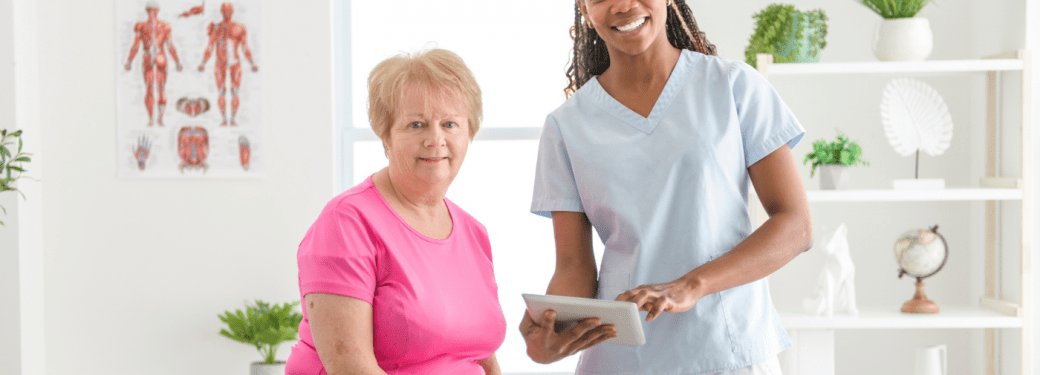 Registered Massage Therapist in scrubs holding tablet next to seated person smiling.