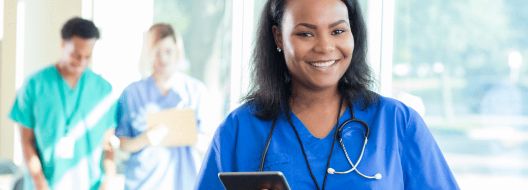 A healthcare professional wearing scrubs and a stethoscope and holding a tablet smiling at the viewer. Two other healthcare workers wearing scrubs are in the background.