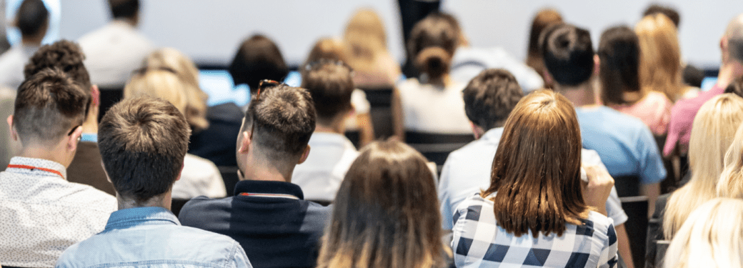 Many people seated listening to a presenter blurred in the background at a conference event.