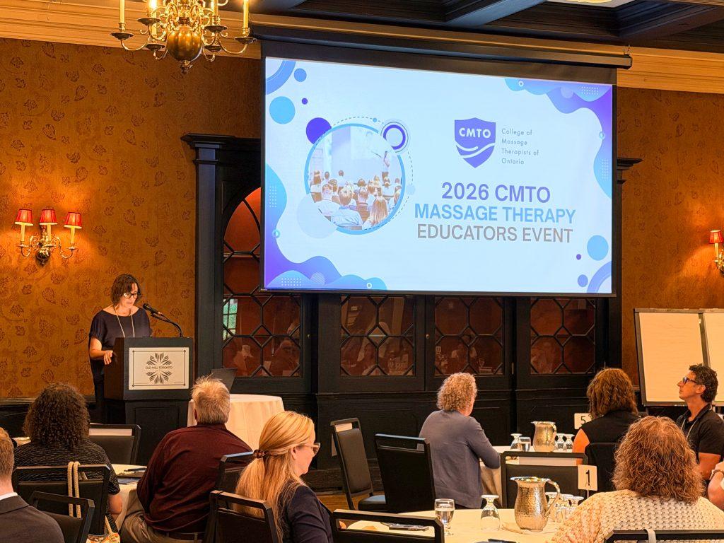 Massage Therapy educators sitting in a room with round tables listening to the College of Massage Therapists of Ontario's Registrar & CEO, Maureen Boon, speak in front of a large project screen with an introduction slide titled "2026 CMTO Massage Therapy Educators Event".