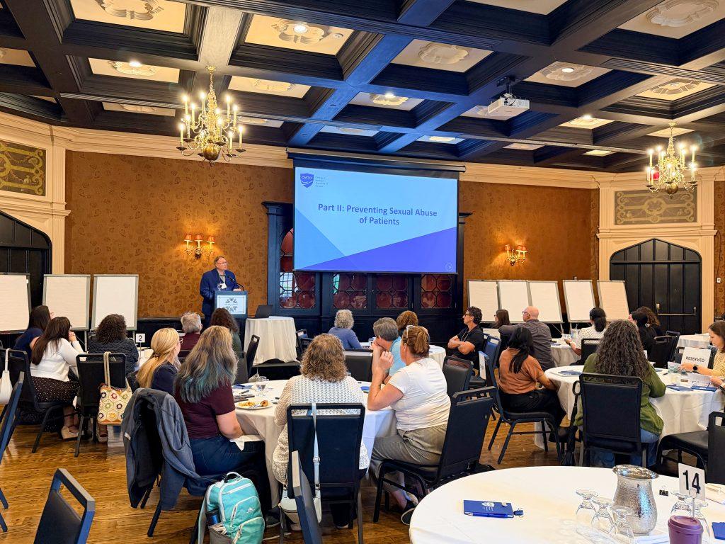 Massage Therapy educators sitting in a room with round tables listening to the College of Massage Therapists of Ontario staff speak in front of a large project screen with an introduction text titled "Part II: Preventing Sexual Abuse of Patients".