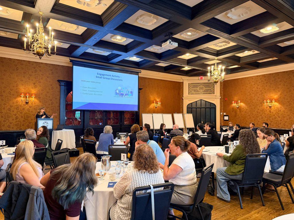 Massage Therapy educators sitting in a room with round tables listening to the College of Massage Therapists of Ontario staff speak in front of a large project screen with a text about engagement activity and small group discussion.