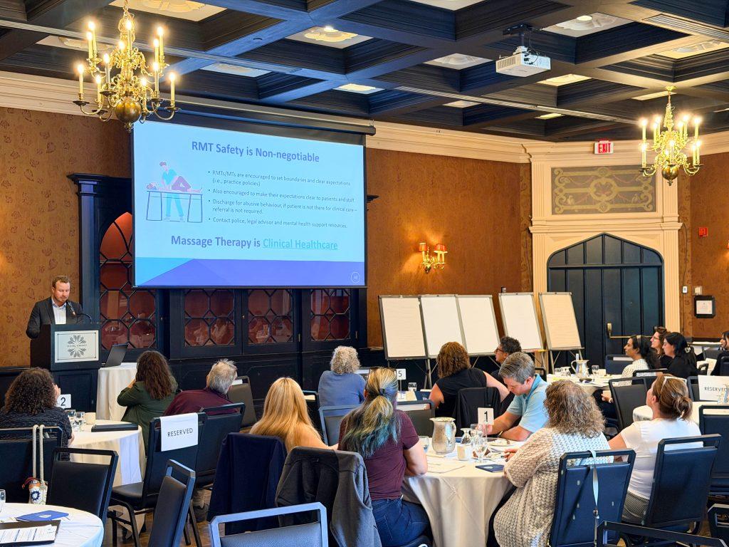 Massage Therapy educators sitting in a room with round tables listening to the College of Massage Therapists of Ontario staff speak in front of a large project screen with a text about the safety of RMTs and that Massage Therapy is clinical healthcare.